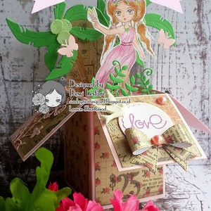 May include: A pink and green papercraft box featuring a fairy and flowers. The box has a patterned paper exterior and a white label with the word "love" on it. The fairy is holding a flower and has a pink dress and brown hair. The box is decorated with green leaves and flowers.