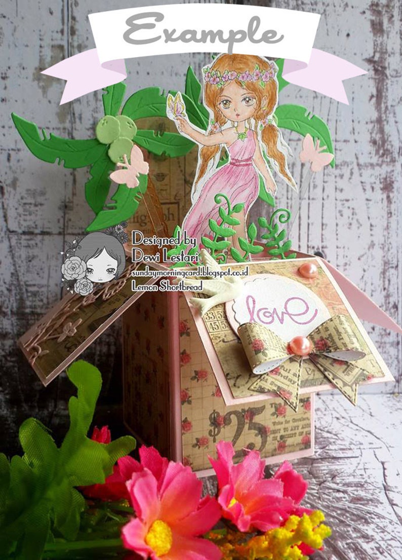 May include: A pink and green papercraft box featuring a fairy and flowers. The box has a patterned paper exterior and a white label with the word "love" on it. The fairy is holding a flower and has a pink dress and brown hair. The box is decorated with green leaves and flowers.