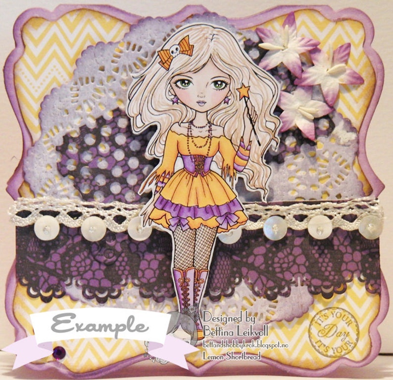 May include: A purple and yellow card with a whimsical illustration of a girl with blonde hair, wearing a yellow dress and holding a wand. The card has a lace border and a scalloped edge. The text "Example" is printed on the card. The text "Designed by Bettina Leikvoll" is printed on the card. The text "It's Your Day It's Your Day" is printed on the card.
