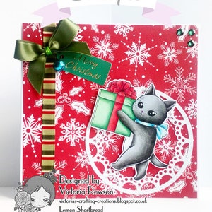 May include: A red Christmas card with a white snowflake pattern. A green ribbon with a bow is tied to the top left corner. A green tag with the text "Merry Christmas" is attached to the ribbon. A black cat with a blue bow tie is holding a green gift box with a red bow. The cat is standing on a white doily. The card is designed by Victoria Rowson and is called "Lemon Shortbread".
