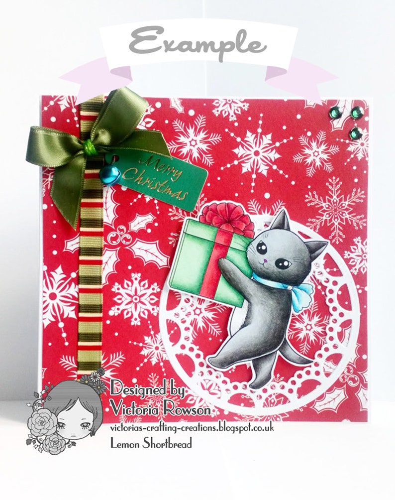 May include: A red Christmas card with a white snowflake pattern. A green ribbon with a bow is tied to the top left corner. A green tag with the text "Merry Christmas" is attached to the ribbon. A black cat with a blue bow tie is holding a green gift box with a red bow. The cat is standing on a white doily. The card is designed by Victoria Rowson and is called "Lemon Shortbread".