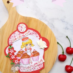 K&ouml;nnte beinhalten: Ein rotes und wei&szlig;es Papierhandwerk mit einem M&auml;dchen, das Cupcakes h&auml;lt und den Text "Keep Calm and Eat Cake". Das Handwerk befindet sich auf einem Holzbrett mit roten Kirschen an der Seite.