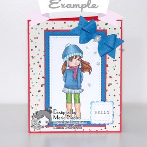May include: A handmade greeting card featuring a girl wearing a blue coat, green pants, and a blue hat. The card has a red, white, and blue patterned background with a blue bow on top. The card says "Designed by Maria Nicoira" and "Lemon Shortbread" on the bottom.
