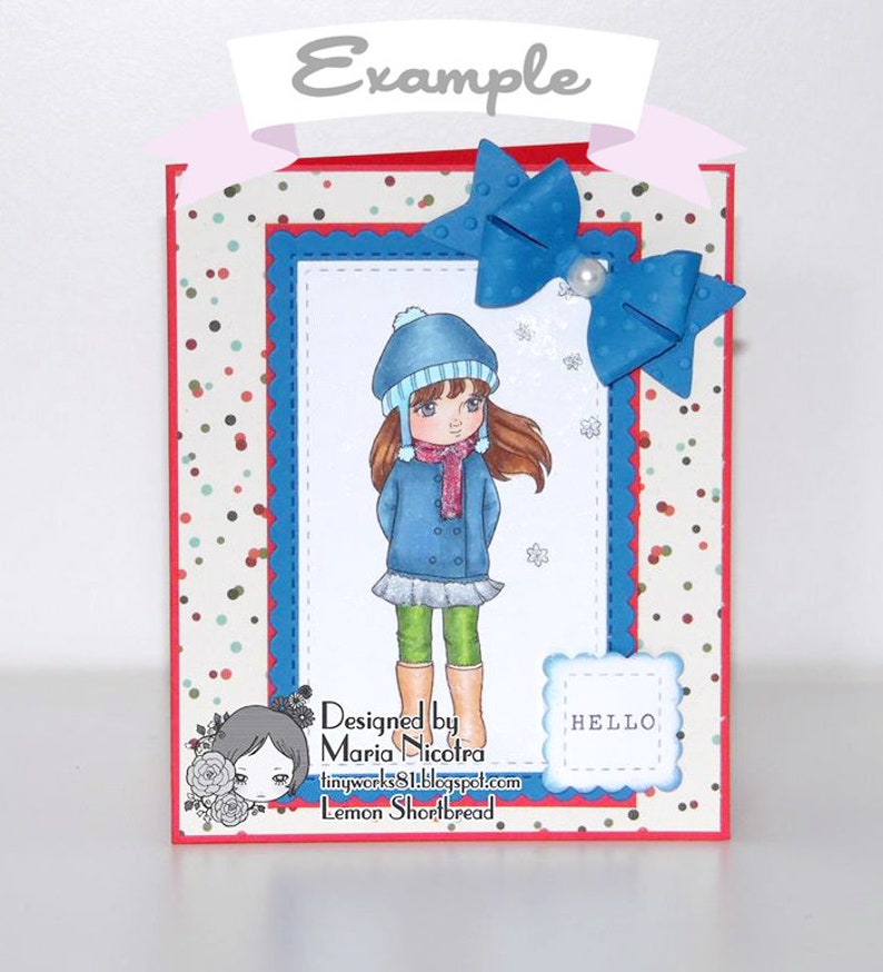 May include: A handmade greeting card featuring a girl wearing a blue coat, green pants, and a blue hat. The card has a red, white, and blue patterned background with a blue bow on top. The card says "Designed by Maria Nicoira" and "Lemon Shortbread" on the bottom.