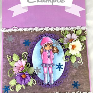 May include: A purple gift bag with a white lace trim and a white ribbon with the word "Example" printed on it. The bag features a brown background with a girl wearing a pink coat and a white hat standing in a purple oval frame. The girl is surrounded by snowflakes and flowers. The bag also has the text "Spiral Creativity" printed on the bottom.
