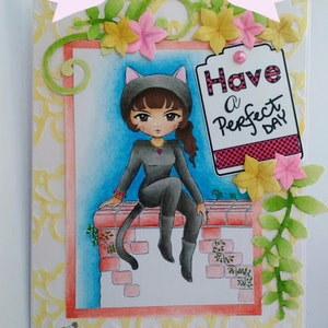 May include: A greeting card with a yellow and pink floral design. The card features a cartoon cat wearing a black catsuit and sitting on a brick wall. The card has the text "Have a Perfect Day" on a pink and white striped tag.