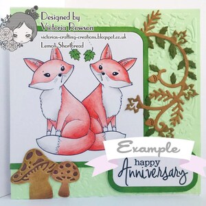 May include: A handmade card with a green background and a white border. The card features two red foxes with white accents, a brown vine with green leaves, and two brown mushrooms. The card also includes the text "Example happy Anniversary".