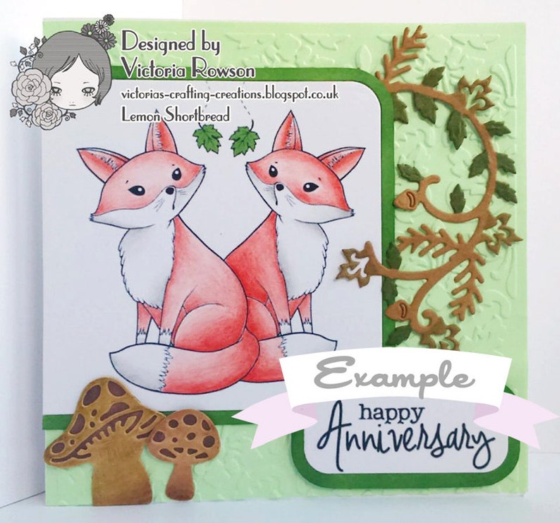 May include: A handmade card with a green background and a white border. The card features two red foxes with white accents, a brown vine with green leaves, and two brown mushrooms. The card also includes the text "Example happy Anniversary".