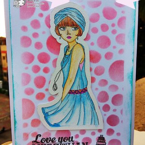 May include: A greeting card with a pink and white polka dot background. A woman with a blue turban and a blue dress is featured on the card. The text "Love you more than" is printed on the card with a heart symbol and a small cake image.