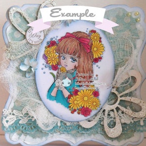 May include: A white card with a blue border and a white oval in the center. The oval contains a drawing of a girl with long brown hair wearing a blue dress and a red bow. She is holding a small doll with blonde hair. The oval is surrounded by flowers and the text "Designed by Bettina Leikvoll bettinahobbykrock.blogspot.ne Lemon Shortbread". The card also has a white ribbon with the text "Example" on it.