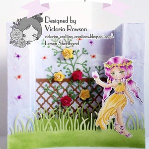 May include: A handmade card featuring a fairy with pink hair and a yellow dress standing in front of a trellis with green vines and red and yellow flowers. The card is designed by Victoria Rowson and is called "Lemon Shortbread".
