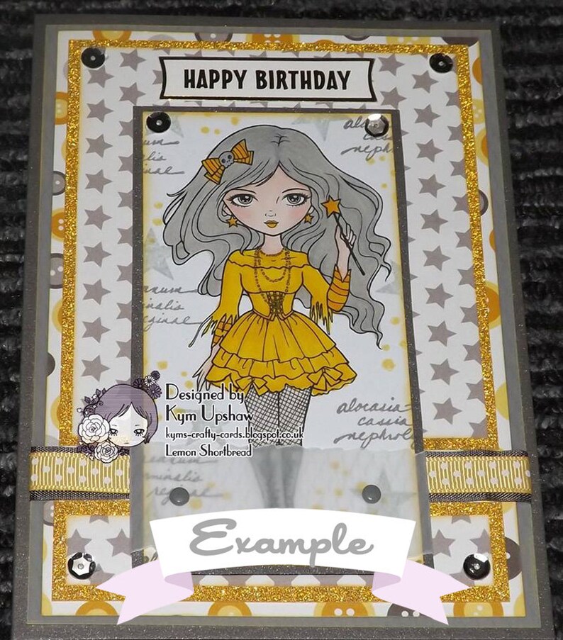 May include: A birthday card with a whimsical illustration of a girl with long gray hair wearing a yellow dress and holding a wand. The card has a gold glitter border and the words "HAPPY BIRTHDAY" at the top. The card also includes the text "Designed by Kym Upshaw" and "Lemon Shortbread".