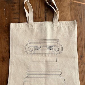May include: A beige canvas tote bag with a black line drawing of an Ionic column. The column design includes a detailed capital with volutes and a fluted base. The bag has two beige handles.