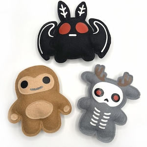 Peut inclure: Trois peluches en feutre faites à la main : un papillon noir avec des yeux rouges, un Bigfoot marron et un cerf gris avec un visage de crâne. Chaque jouet a des accents blancs sur fond blanc.