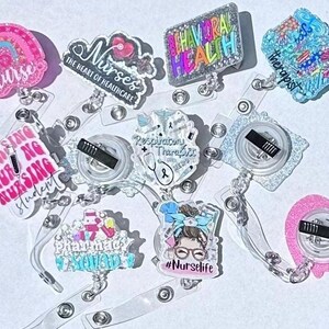 Op de afbeelding: Een verzameling intrekbare badge reels met verschillende ontwerpen en tekst. De reels tonen zinnen als "Nurse", "Nursing Student" en "Behavioral Health". De ontwerpen omvatten regenbogen, harten en medische symbolen. De reels zijn transparant met een metalen clip.