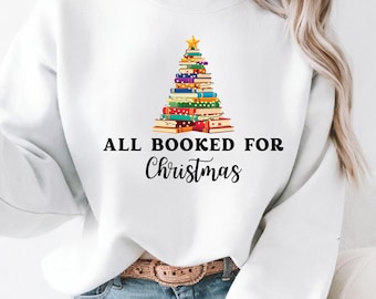 以下が含まれることがあります： 「ALL BOOKED FOR Christmas」の文字が入った白いスウェットシャツ。カラフルな本で作られたクリスマスツリーのグラフィックで、金色の星が飾られています。モデルは青いジーンズと茶色のベルトを着用しています。
