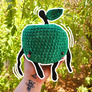 May include: A green crocheted apple plush toy with black legs and a green leaf on top. The toy is being held in a person's hand.
