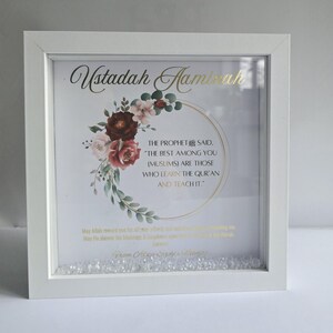 May include: White framed artwork with gold text reading "Ustadah Haminah." The artwork includes a floral design with burgundy and pink roses. The text "THE PROPHET SAID, 'THE BEST AMONG YOU (MUSLIMS) ARE THOSE WHO LEARN THE QUR'AN AND TEACH IT."
