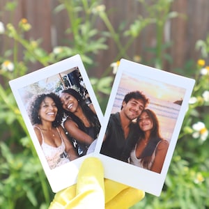 May include: Two white-bordered instant film photographs. The left photo shows two smiling individuals with dark hair. The right photo shows a couple posing on a beach at sunset. The prints are held against a background of green foliage and white flowers.