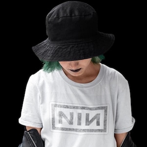May include: A white t-shirt featuring the letters "NIN" within a rectangular border. The outfit includes a black bucket hat and a black leather jacket. The background is a plain black.