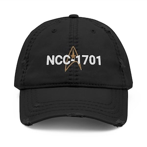 Puede incluir: Gorra de béisbol negra desgastada con el texto "NCC-1701" en blanco y un emblema dorado. La gorra tiene una visera curva y un aspecto desgastado, lo que sugiere un estilo vintage o retro.