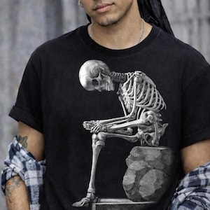 Może przedstawiać: Czarny t-shirt z szarą ilustracją siedzącego szkieletu. Szkielet przedstawiony jest ze spuszczoną głową, spoczywającą na kamieniu. Projekt jest wyśrodkowany na koszulce, tworząc uderzający efekt wizualny.
