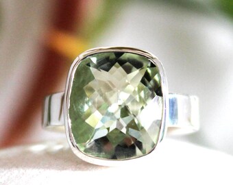 May include: A silver ring featuring a large, square-cut, pale green gemstone. The gemstone has multiple facets, reflecting light. The ring's band is a simple, polished silver. The setting is a bezel style, encasing the stone.