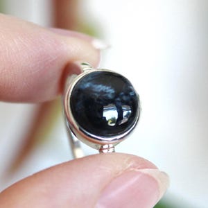May include: A close-up of a silver ring featuring a large, round, polished black gemstone with blue and white marbling. The ring is held between two fingers against a soft, blurred background.