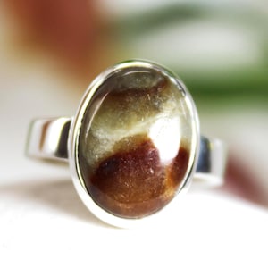 Op de afbeelding: Een zilveren ring met een ovale edelsteen in bruin, beige en witte tinten. De edelsteen is gezet in een gepolijste zilveren band. Dit sieraad heeft een eenvoudig, elegant ontwerp. De ring is een sieraad.