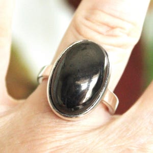 May include: A silver ring featuring a large, oval-shaped, polished black stone. The stone is set in a simple, elegant silver band. The ring is shown on a person's finger, with a soft, blurred background.