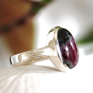 Peut inclure: Une bague en argent ornée d'une pierre précieuse ovale. La pierre affiche une teinte rouge profond avec des inclusions vert foncé. Le corps de la bague est en argent poli.