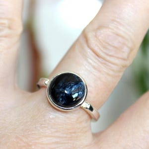 May include: A silver ring featuring a large, round, dark blue gemstone with intricate patterns. The gemstone is set in a simple, polished silver band. The ring is shown on a finger.