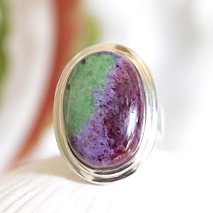 May include: Oval-shaped ring featuring a large, polished gemstone. The stone displays a striking color contrast, with a vibrant green section on one side and a deep purple-red hue on the other. The gemstone is set in a silver-colored metal band.