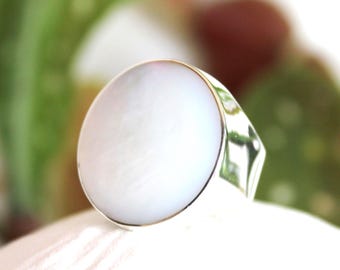 May include: A silver ring with a large, oval, white mother-of-pearl stone. The ring has a polished silver band and a bezel setting that holds the stone securely. The ring is set against a soft, white background.