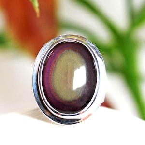 May include: Oval-shaped ring with a silver band. The ring features a large, polished stone with a color gradient of purple, red, and yellow. The stone is set in a bezel setting, creating a modern and elegant look. A unique piece of jewelry.