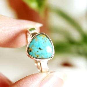 May include: A silver ring featuring a triangular turquoise stone. The stone is a vibrant blue-green color with brown and black inclusions. The ring is held between fingers, showcasing the polished metal band and bezel setting.