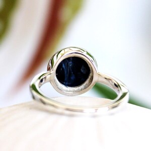 May include: A silver ring with a round, dark blue gemstone. The gemstone has a swirling pattern. The ring is set on a white surface, with a blurred background of green and white.