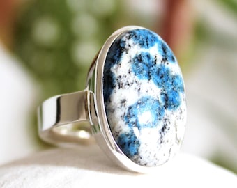 May include: A silver ring featuring a large, oval-shaped k2 stone. The stone has a striking pattern of blue, white, and black, set in a polished silver band. The ring's design is simple, highlighting the natural beauty of the stone.