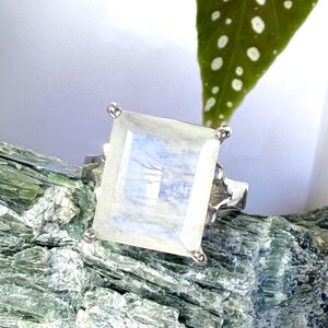 May include: A silver ring featuring a large, rectangular, faceted moonstone with a translucent, milky white appearance and subtle blue iridescence. The gemstone is held in place by four silver prongs on a polished silver band with decorative side details.