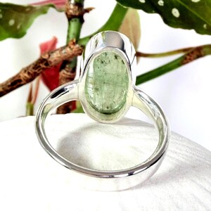 May include: A close-up of a silver ring featuring a large, oval-shaped, pale green gemstone. The ring's band is smooth and polished, and the gemstone is set in a bezel setting. The ring is displayed against a white background with green foliage.