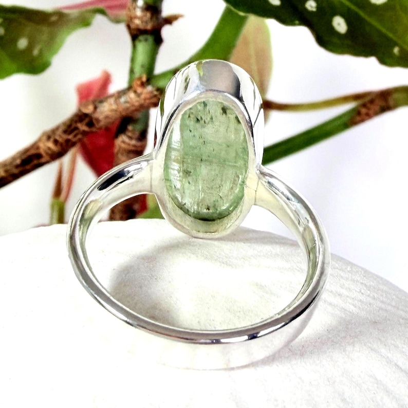 May include: A close-up of a silver ring featuring a large, oval-shaped, pale green gemstone. The ring's band is smooth and polished, and the gemstone is set in a bezel setting. The ring is displayed against a white background with green foliage.