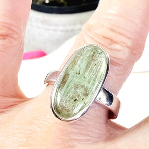 May include: A silver ring featuring an oval-shaped, light green gemstone. The gemstone has a textured appearance with darker green inclusions. The ring's band is a simple, polished silver design.