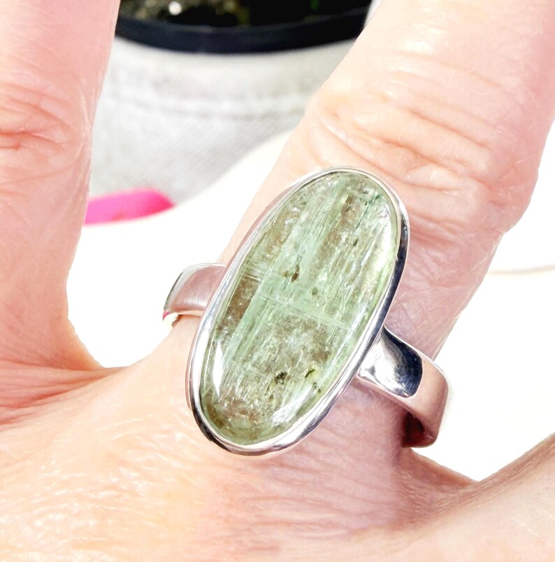 May include: A silver ring featuring an oval-shaped, light green gemstone. The gemstone has a textured appearance with darker green inclusions. The ring's band is a simple, polished silver design.