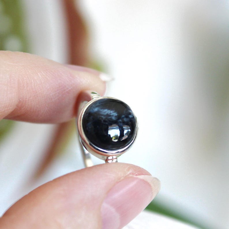 May include: A close-up of a silver ring featuring a large, round, polished black gemstone with blue and white marbling. The ring is held between two fingers against a soft, blurred background.