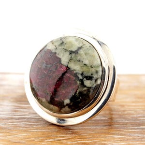 May include: A large, round gemstone ring with a silver band. The gemstone features a mix of colors, including deep red and green, set in a polished silver bezel. The ring is displayed on a wooden surface.