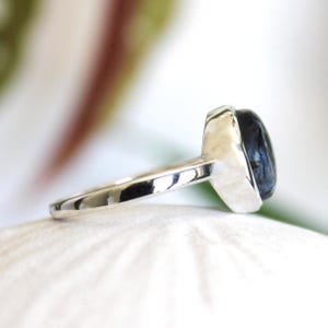 May include: A silver ring with a dark blue, oval-shaped gemstone. The ring's band is smooth and polished, leading up to the setting that holds the stone. The gemstone has a polished, reflective surface.