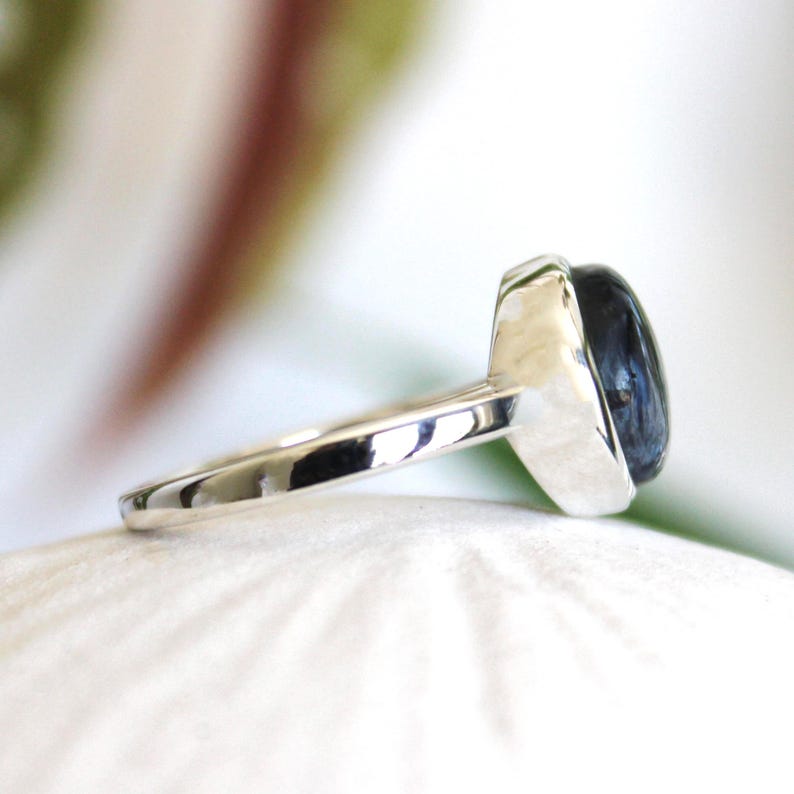 May include: A silver ring with a dark blue, oval-shaped gemstone. The ring's band is smooth and polished, leading up to the setting that holds the stone. The gemstone has a polished, reflective surface.
