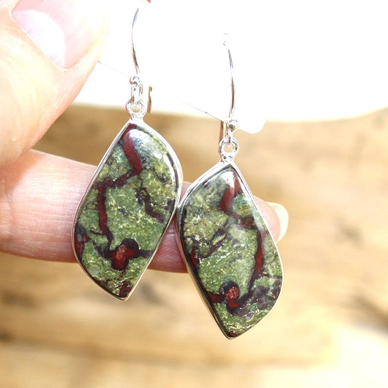 May include: A pair of dangle earrings featuring green and red patterned stones set in silver-colored metal. The earrings have a teardrop shape and are suspended from a simple hook.