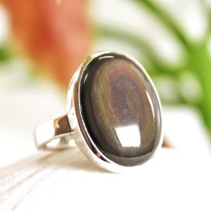 May include: A silver ring featuring a large, oval-shaped gemstone with a dark, iridescent sheen. The stone displays a mix of colors, including brown, green, and gold. The ring's band is a simple, polished silver.