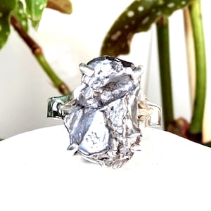 May include: A silver ring featuring a large, irregular, textured metallic centrepiece with a rough, cratered surface. The central element is secured by multiple small prongs extending from a smooth, polished silver band that wraps around the finger.